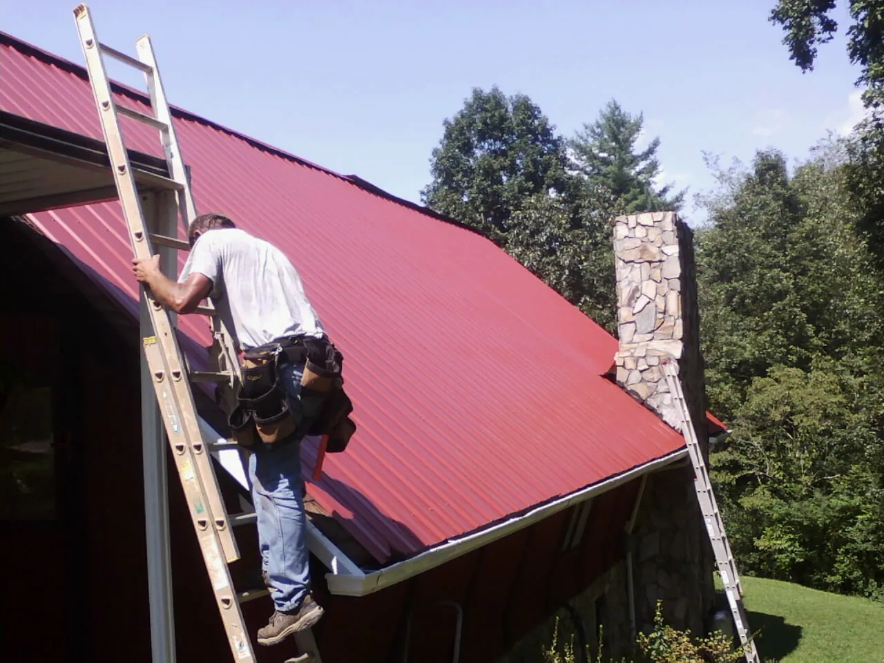 Roofer on a ladder beside a red metal roof and stone chimney — Soffit & Fascia Repair by Roofing Brothers in Battery Park